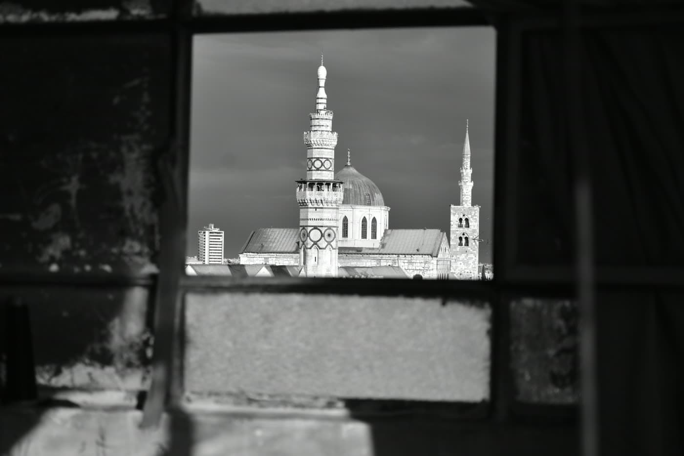 The dome and minarets of the Umayyad Mosque in Damascus, seen in black and white through the frame of a derelict window